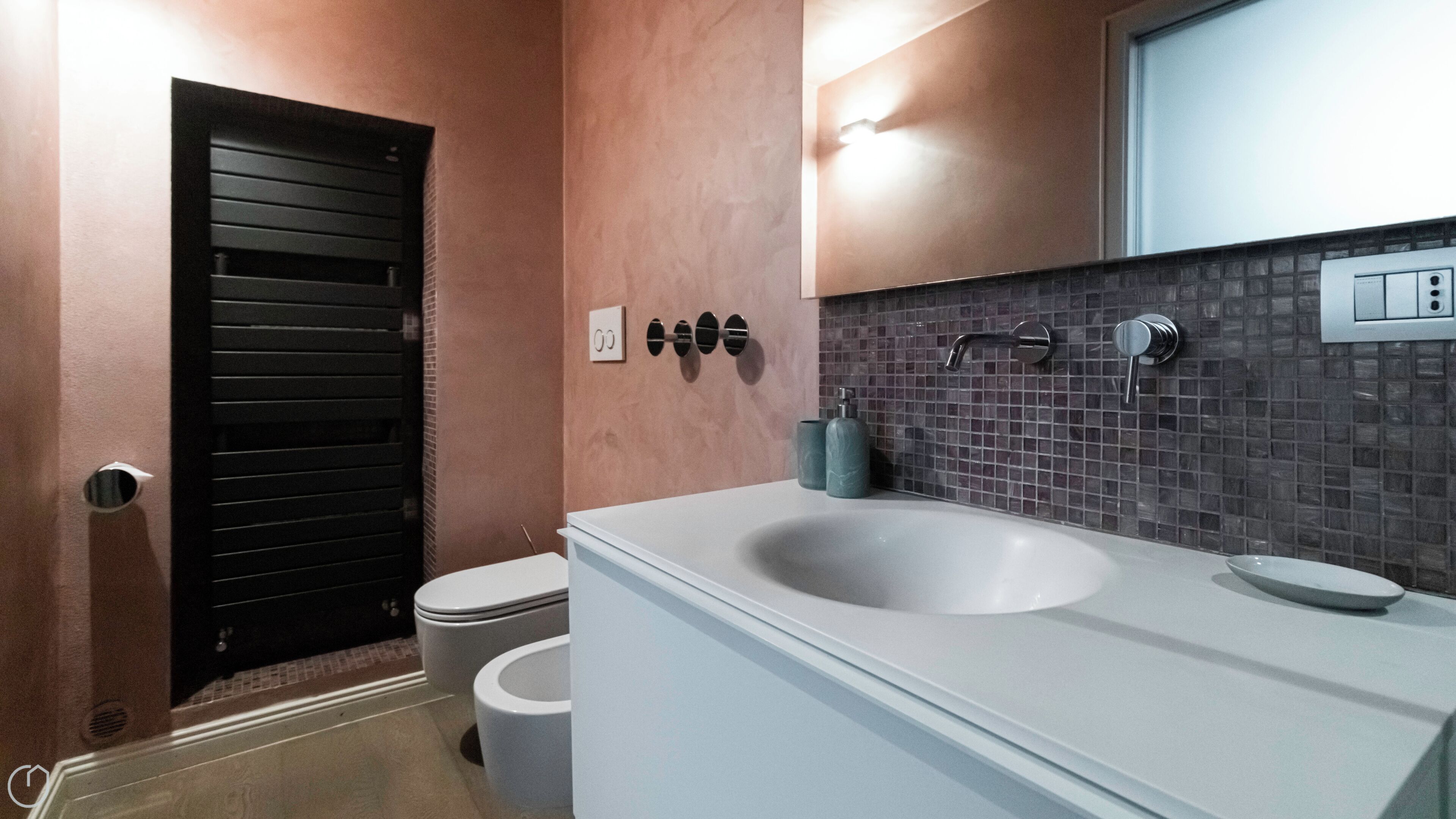 Ein modernes Badezimmer mit einem weißen Waschbecken, einer dunkel gefliesten Rückwand und einer schwarzen Tür.