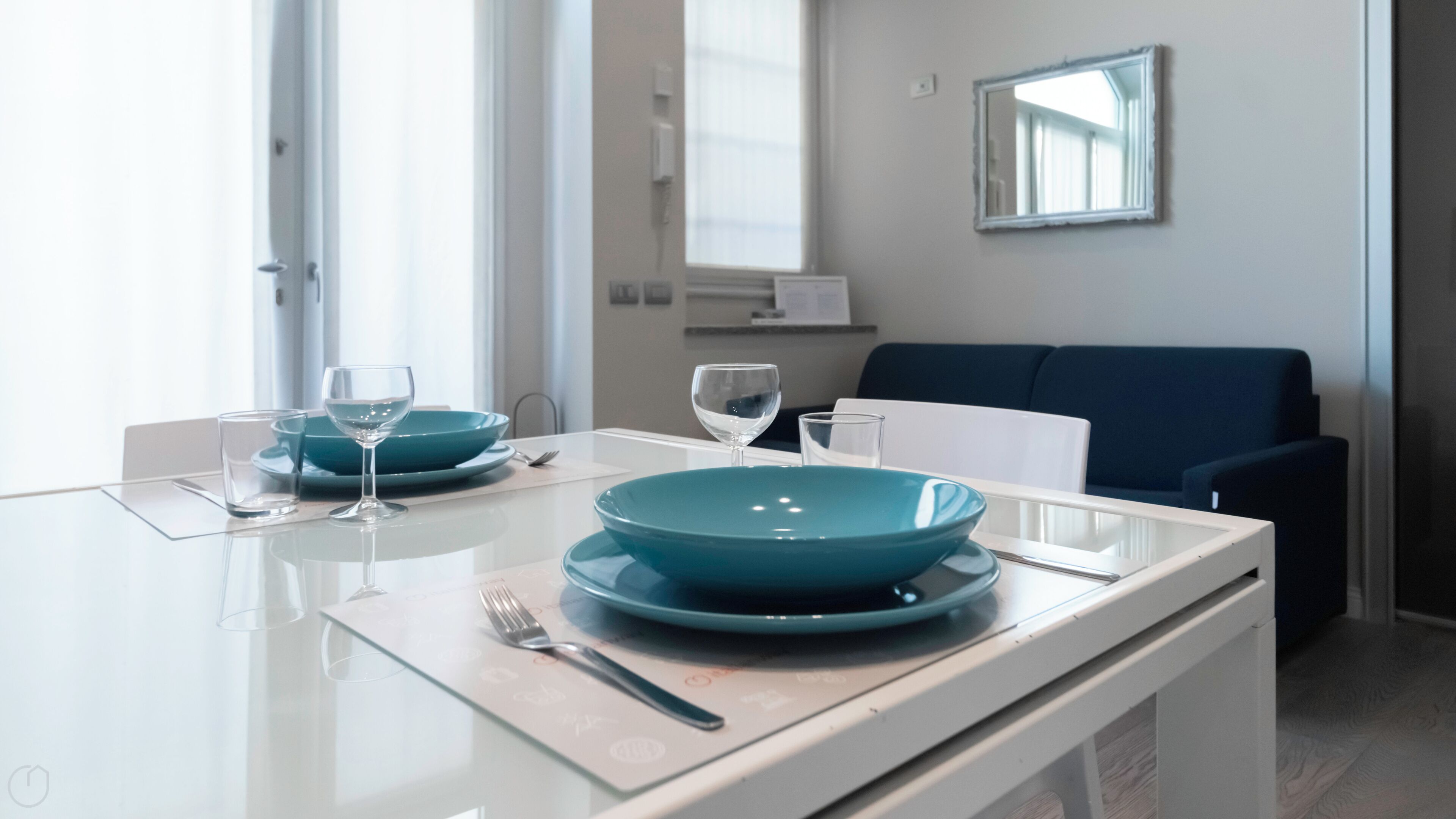 Un comedor moderno con una mesa blanca dispuesta para dos personas, con platos verde agua y copas de cristal.