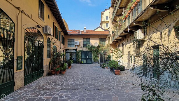 A narrow cobblestone street flanked by buildings with balconies, potted plants, and a central door. - Italianway Easy - Naviglio Pavese 6 (Milan)