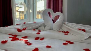 Ein Hotelzimmer mit einem Bett, das mit weißen Leinen und roten Rosenblättern dekoriert ist, einem Fenster mit roten Vorhängen und Blick auf Gebäude.