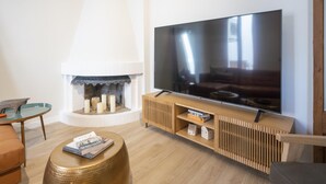 A modern living room with a large flat-screen TV mounted on a wooden console, a fireplace with candles, a round gold table, and a brown leather sofa.