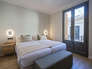 A bedroom with a bed, bedside tables, a bench, and a large window with a view of the outside. - Sitges Spaces Urban Oasis | 3BR/2BA 🌴 (Sitges)