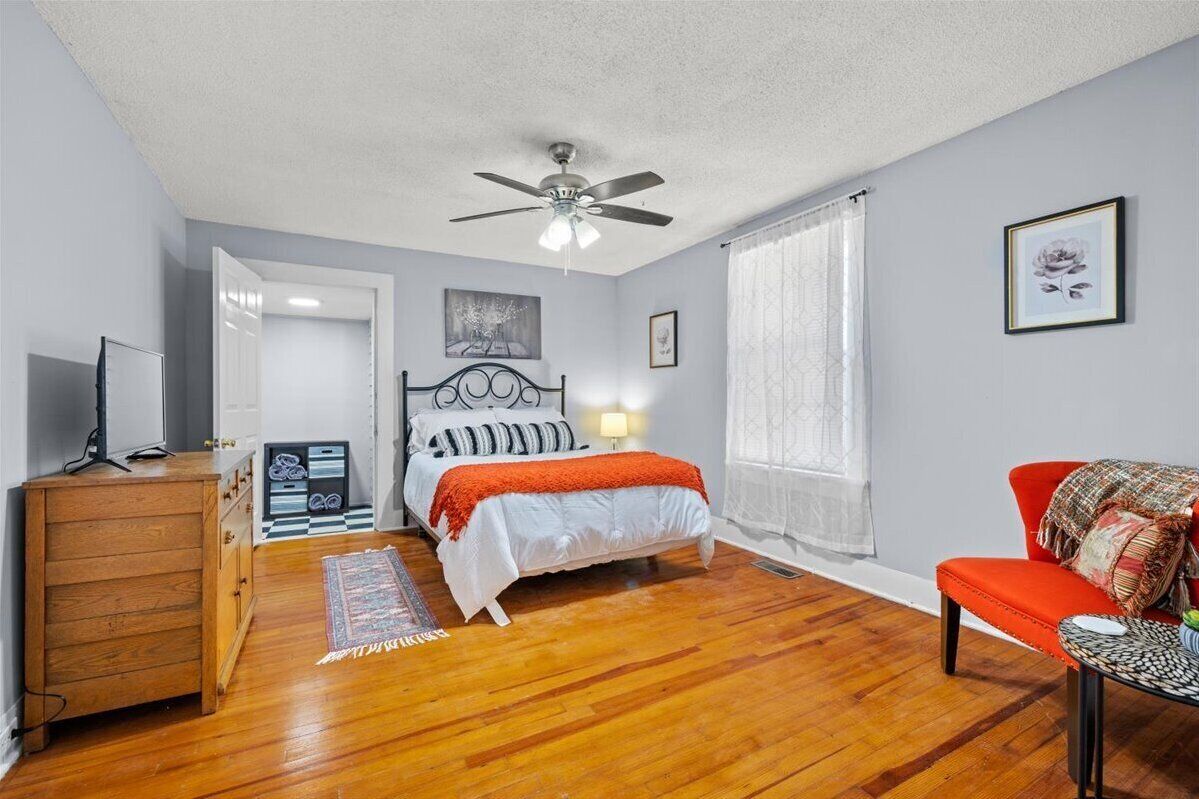 Un dormitorio con cama, ventilador de techo, cómoda de madera, una silla roja y ventana con cortinas.