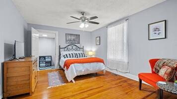 Un dormitorio con cama, ventilador de techo, cómoda de madera, una silla roja y ventana con cortinas.
