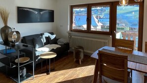 A modern living room with a black sofa, a dining table with chairs, a large window with a view of a town, and a ceiling light fixture. - Family-friendly temporary home (Horben)