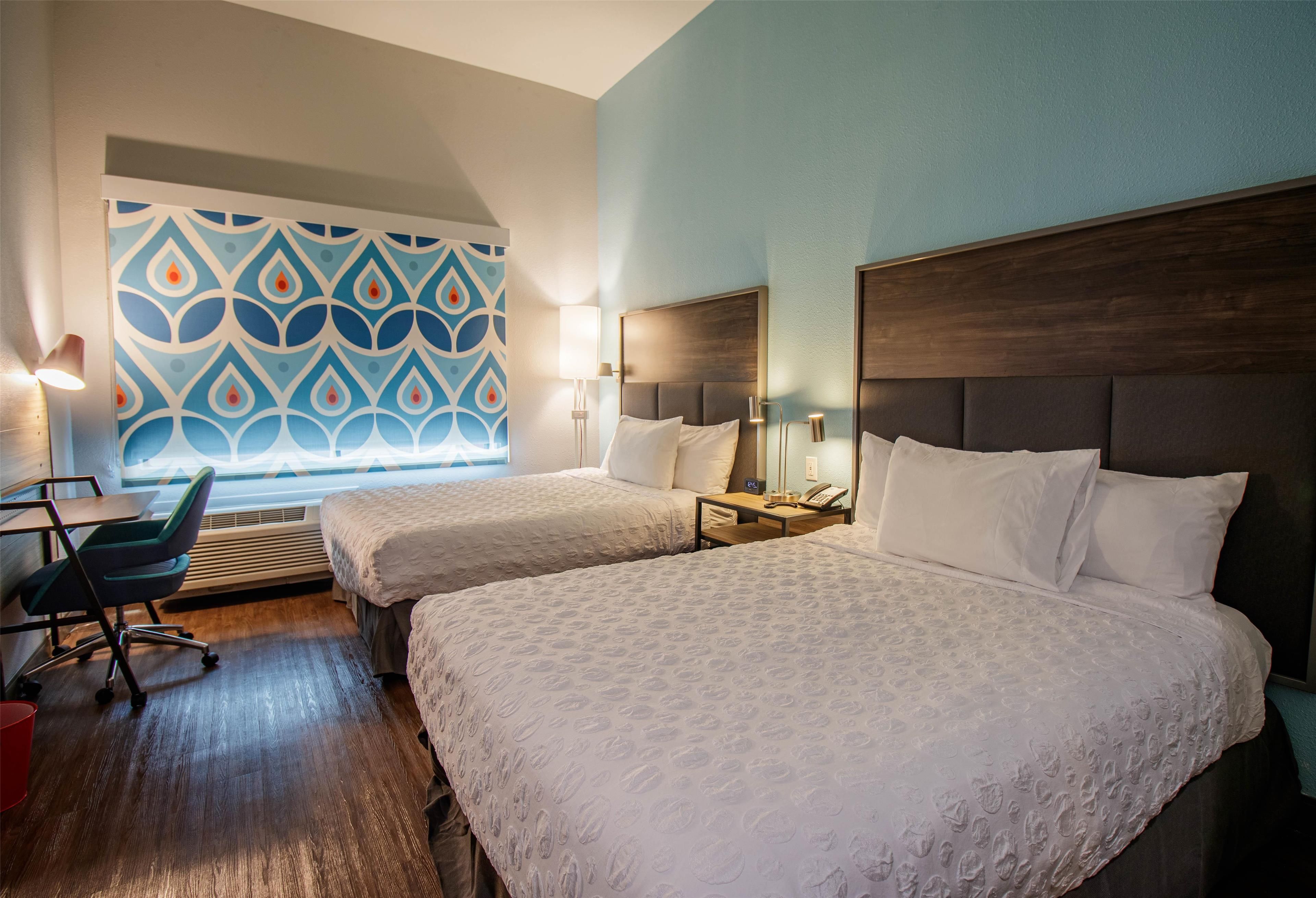 A hotel room with two beds, a desk, a chair, and a patterned wall.