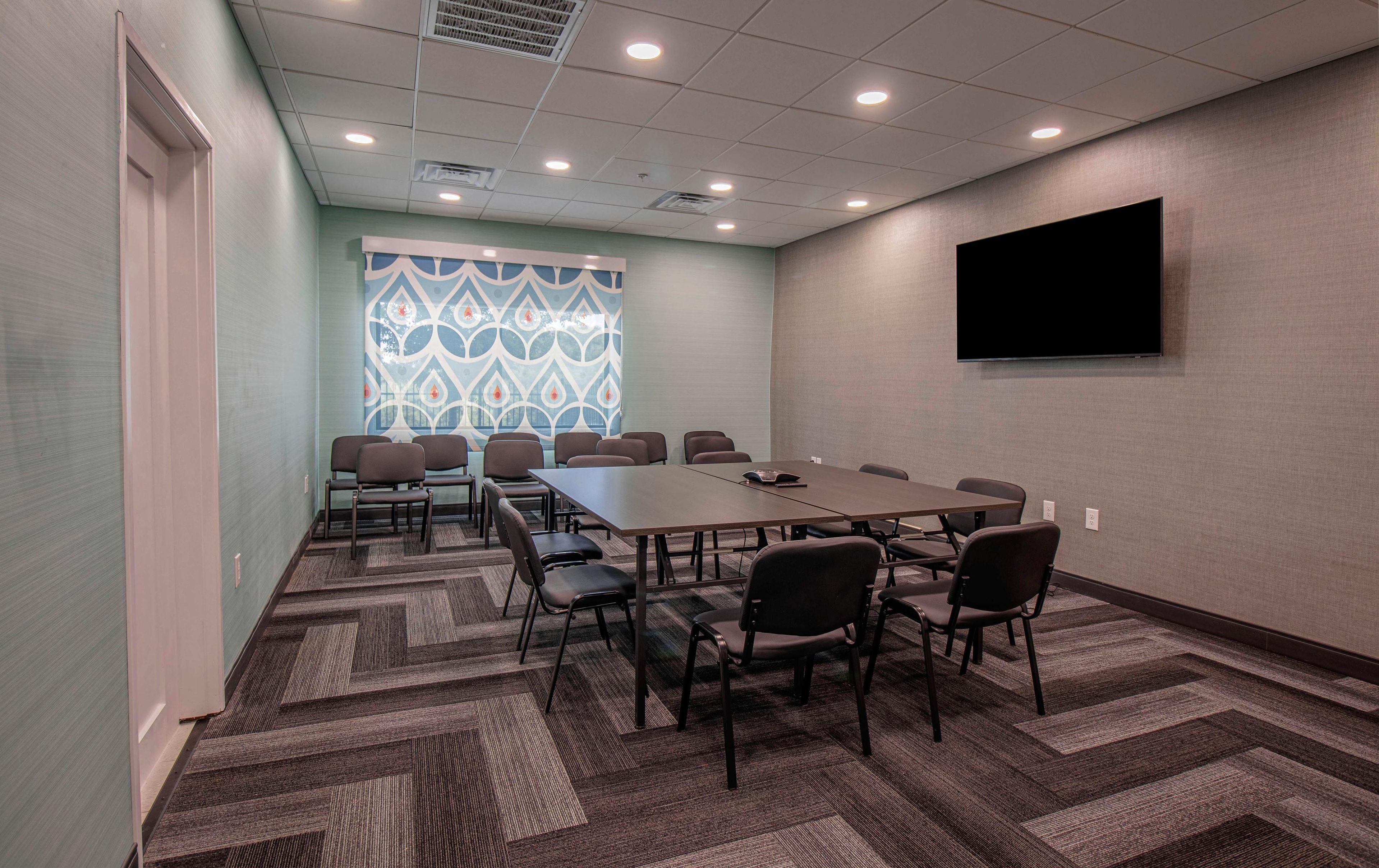 A conference room with a long table, multiple chairs, carpeted flooring, a patterned window, and a mounted TV.