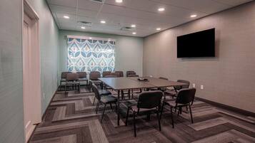 A conference room with a long table, multiple chairs, carpeted flooring, a patterned window, and a mounted TV.