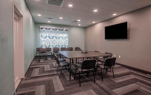 Un salón de conferencias con una larga mesa, varias sillas, piso alfombrado, una ventana con diseño y un televisor montado.