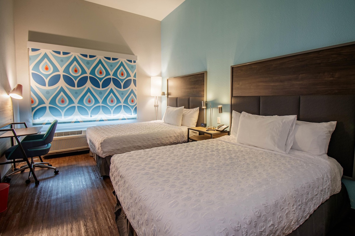 A hotel room with two beds, a desk, a chair, and a patterned wall.