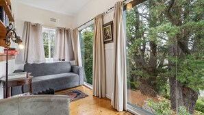 Uma sala de estar aconchegante com um sofá cinza, uma mesinha com livros e vista para uma floresta através de grandes janelas.