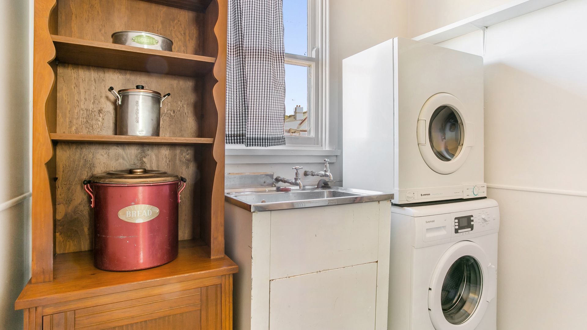 A compact kitchen with a wooden shelf, a red pot, a stainless steel pot, a sink, and a front-loading washing machine.
