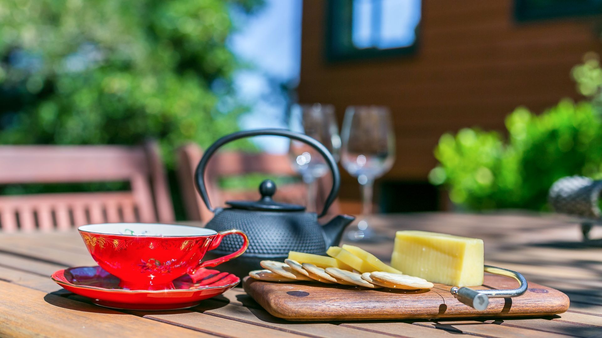 A wooden table with a red teacup, a black teapot, cheese, crackers, and a cheese knife.