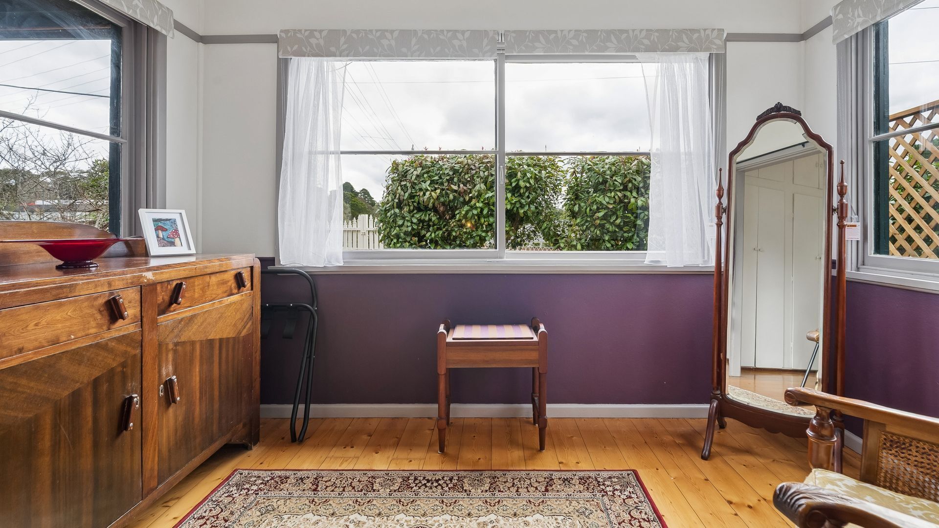 A room with a wooden cabinet, a small wooden stool, a mirror, and a window with a view of greenery.