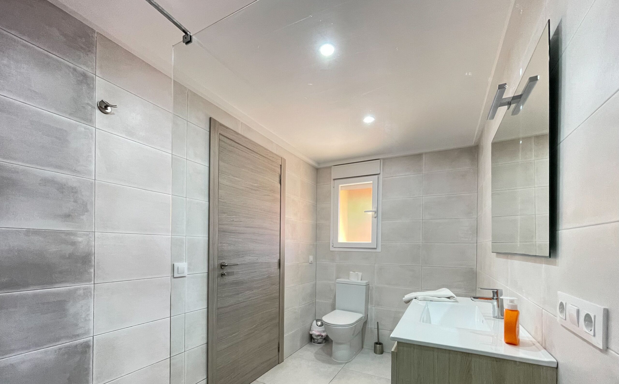 A modern bathroom with grey tiles, a wooden door, a toilet, a sink with a mirror, and a window with daylight.