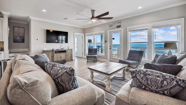A spacious living room with a large sofa set, a coffee table, and a television. There is a ceiling fan, a kitchen area, and a view of the ocean through large windows.