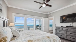 A bedroom with a large bed, a ceiling fan, a TV, and a view of the ocean through the sliding doors.