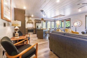 A modern kitchen with wooden cabinets, a dining area with a table and chairs, and a living room with a sofa and a clock on the wall. - BEAR HAVEN - INDOOR BLACK-LIGHT MINI GOLF! (Gatlinburg)
