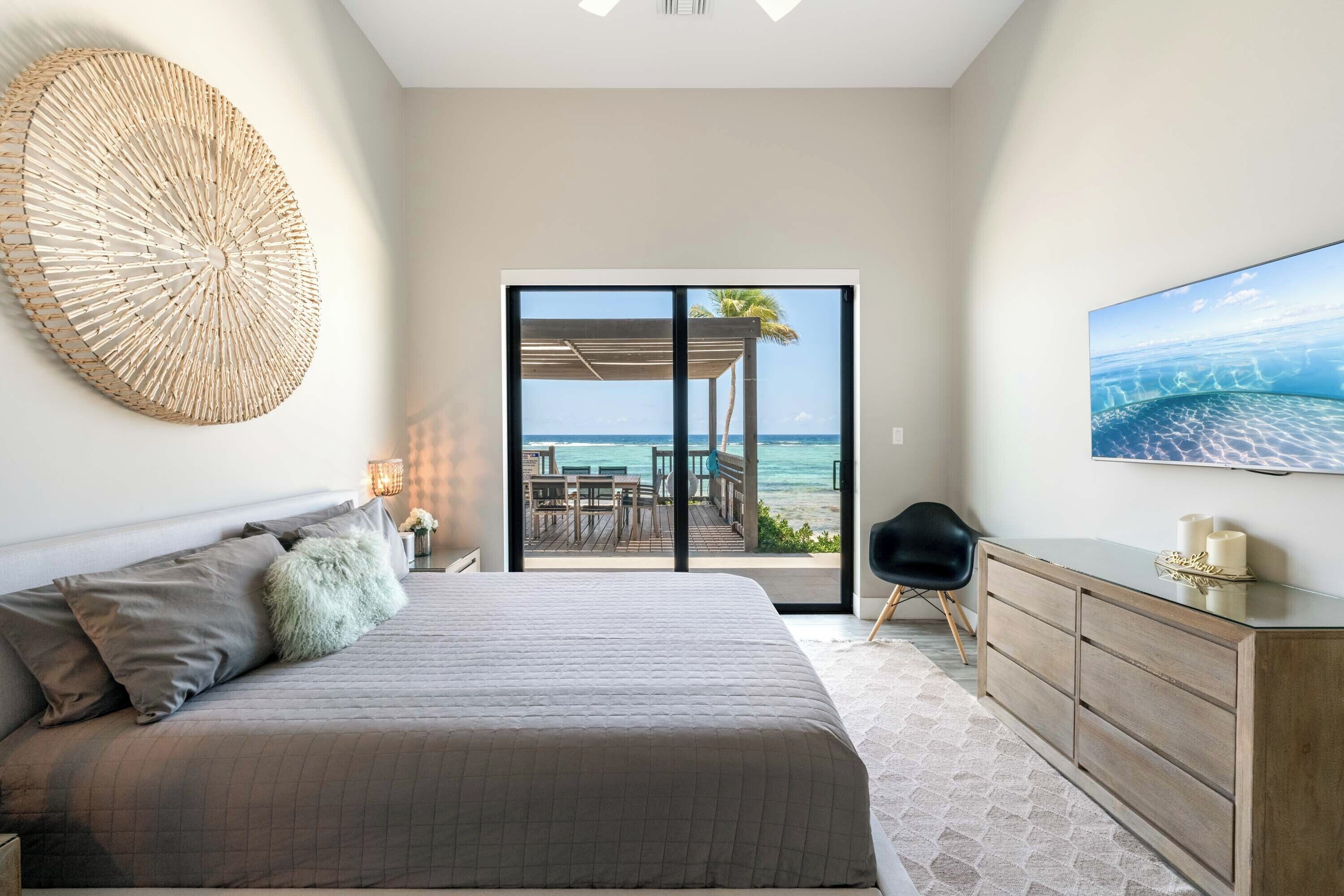 A bedroom with a bed, a dresser, a TV, and a view of the beach through a sliding door.