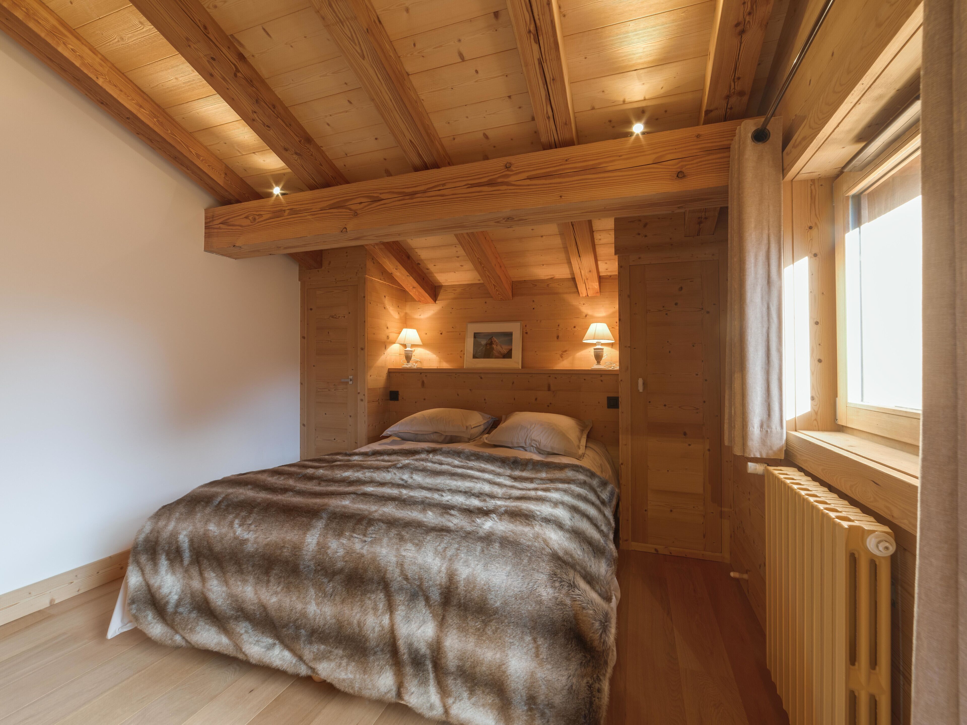 A cozy wooden bedroom with a bed, two bedside lamps, a window with curtains, and a radiator.