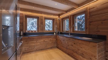 Une cuisine moderne avec des meubles en bois, un réfrigérateur en acier inoxydable et de grandes fenêtres offrant une vue sur les arbres couverts de neige.