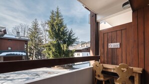 Un balcon avec un banc et des chaises en bois, une vue sur les arbres et les bâtiments, et une pancarte sur le mur.