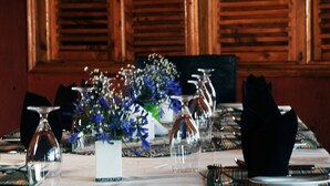 Et spisebord dækket med klare glas, en vase med blå blomster og sorte servietter.