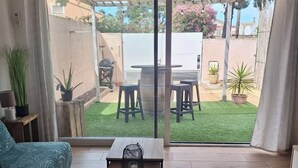 Un salon moderne comprenant un canapé, une table basse et une porte-fenêtre coulissante donnant sur un jardin avec un barbecue et un espace salon extérieur.