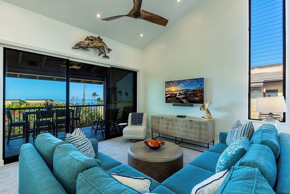 A modern living room with a large sofa, a coffee table, and a TV. There is a balcony with a view of the ocean.