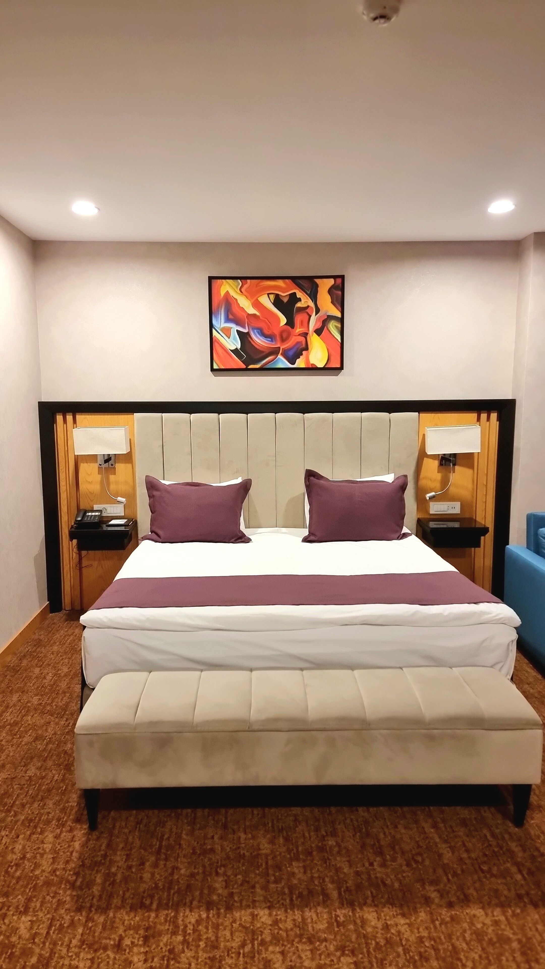 Een hotelkamer met een groot bed, twee nachtkastjes, een hoofdbord met een schilderij en een bankje.