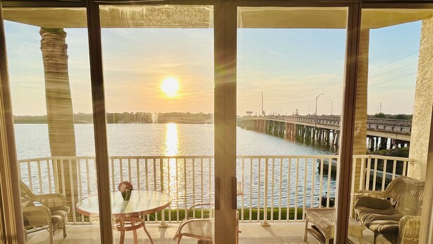 A balcony with a view of a sunset over water, a pier, and outdoor furniture.