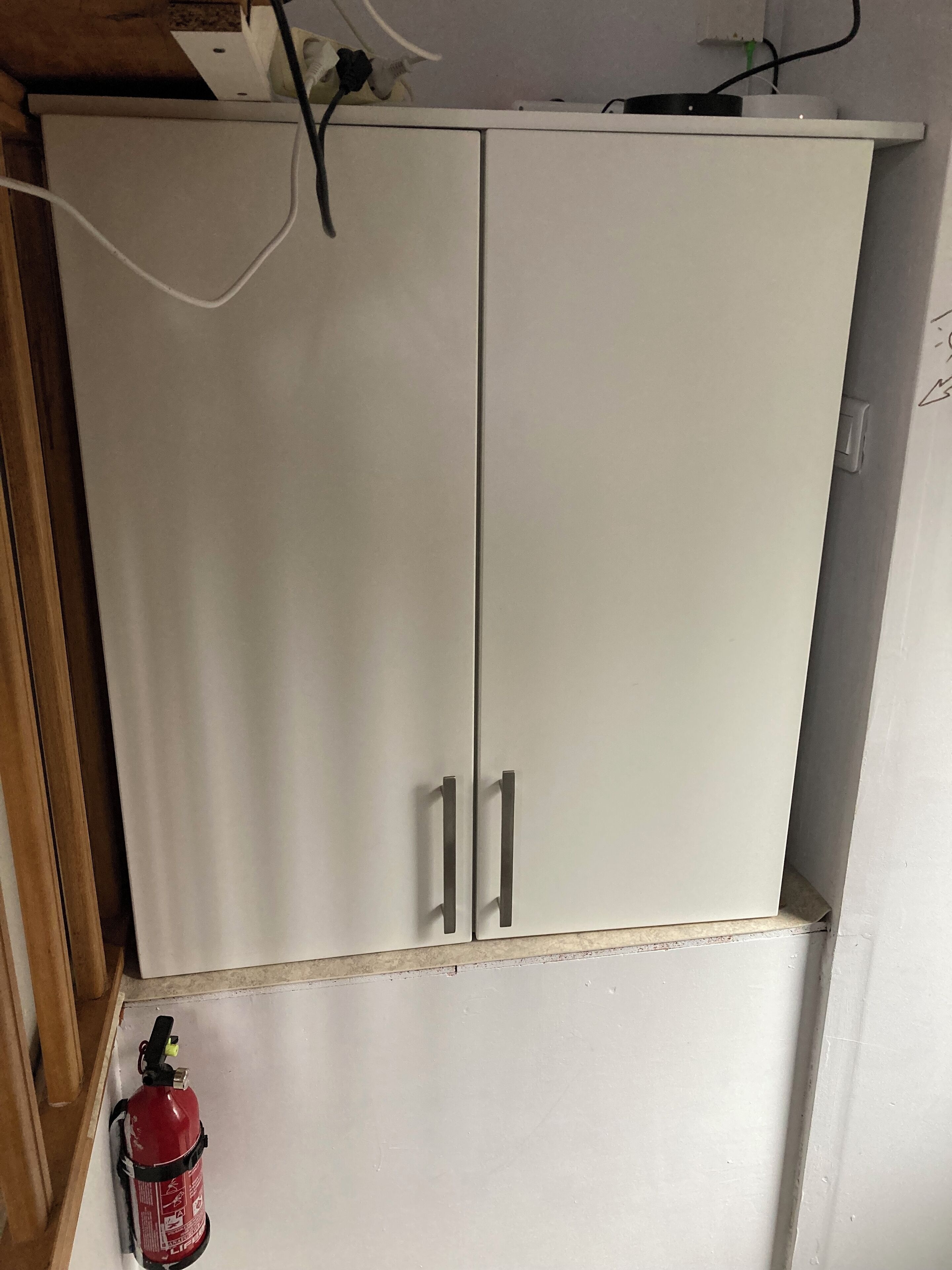 Une armoire blanche à deux portes, avec un extincteur posé sur le sol.