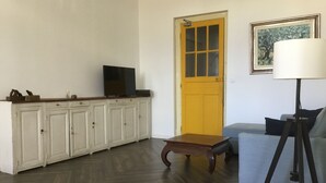 Un salón con suelo de madera en espiga, un armario blanco con un televisor encima, una puerta amarilla, un sofá azul y una lámpara de pie.