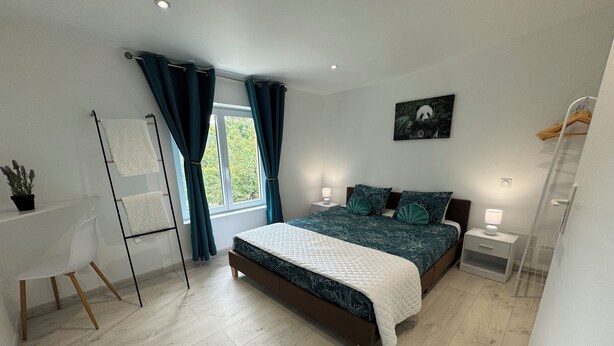 Une chambre moderne avec un grand lit, une table de chevet avec une lampe, une chaise, un bureau avec une plante et une fenêtre avec des rideaux.