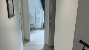 Un couloir aux murs blancs et au plancher en bois. Une porte se trouve sur la droite et, au loin, on aperçoit une chaise blanche.
