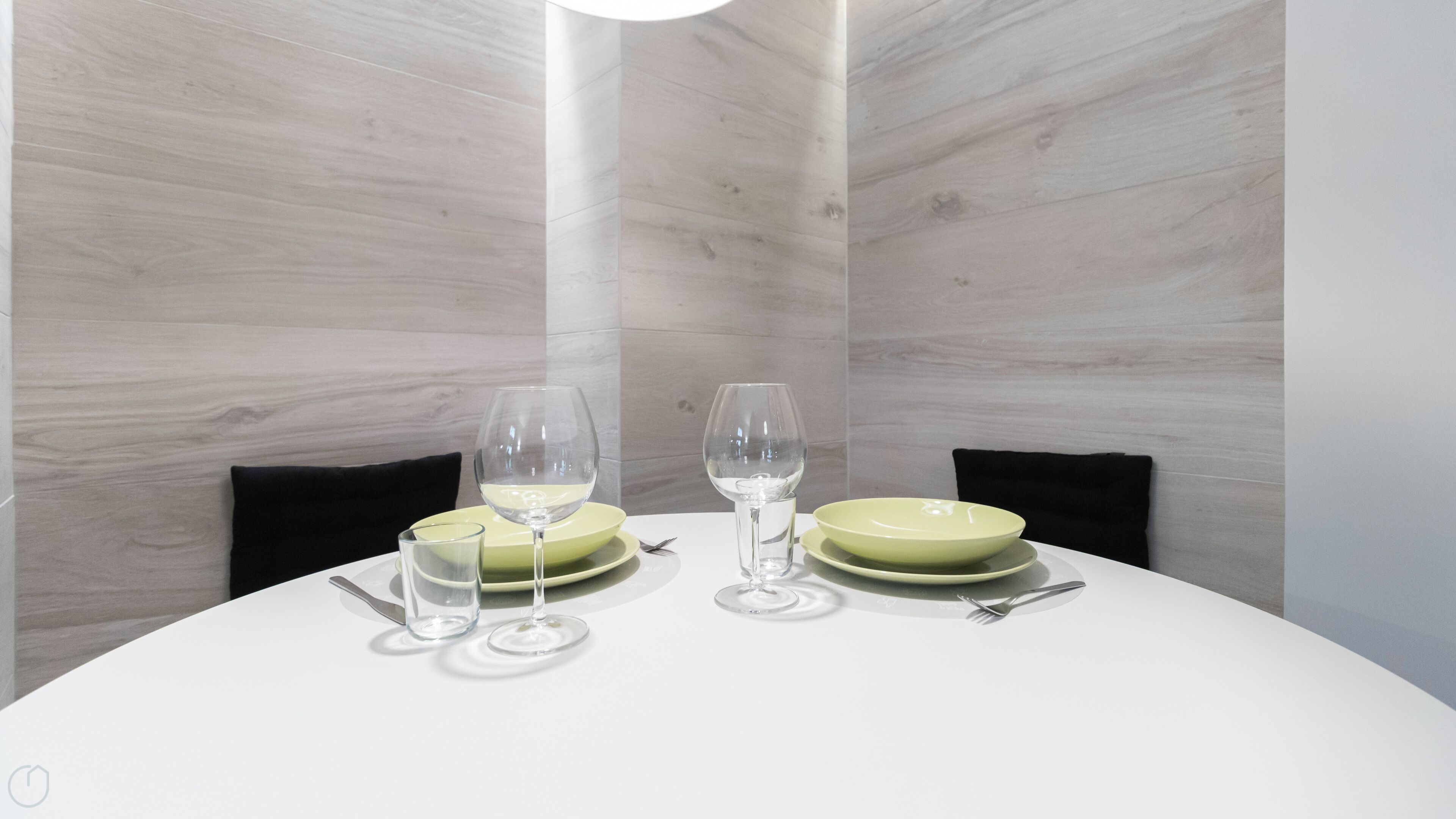 A dining table with two green plates, two wine glasses, and cutlery. The table is set against a wall with a light wood finish.