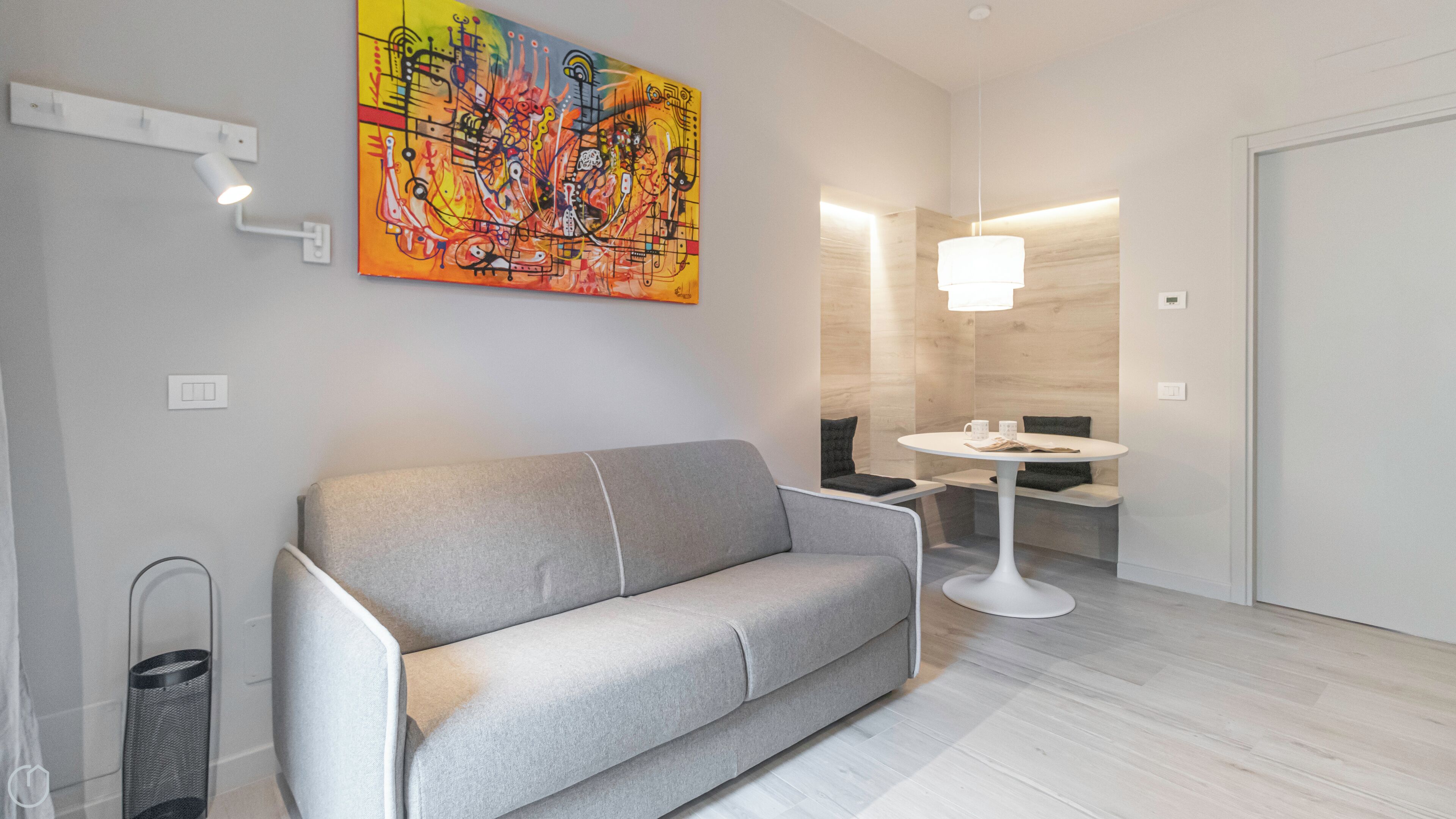 A modern living room with a grey sofa, a colorful abstract painting, a round table with two cups, and a pendant light.