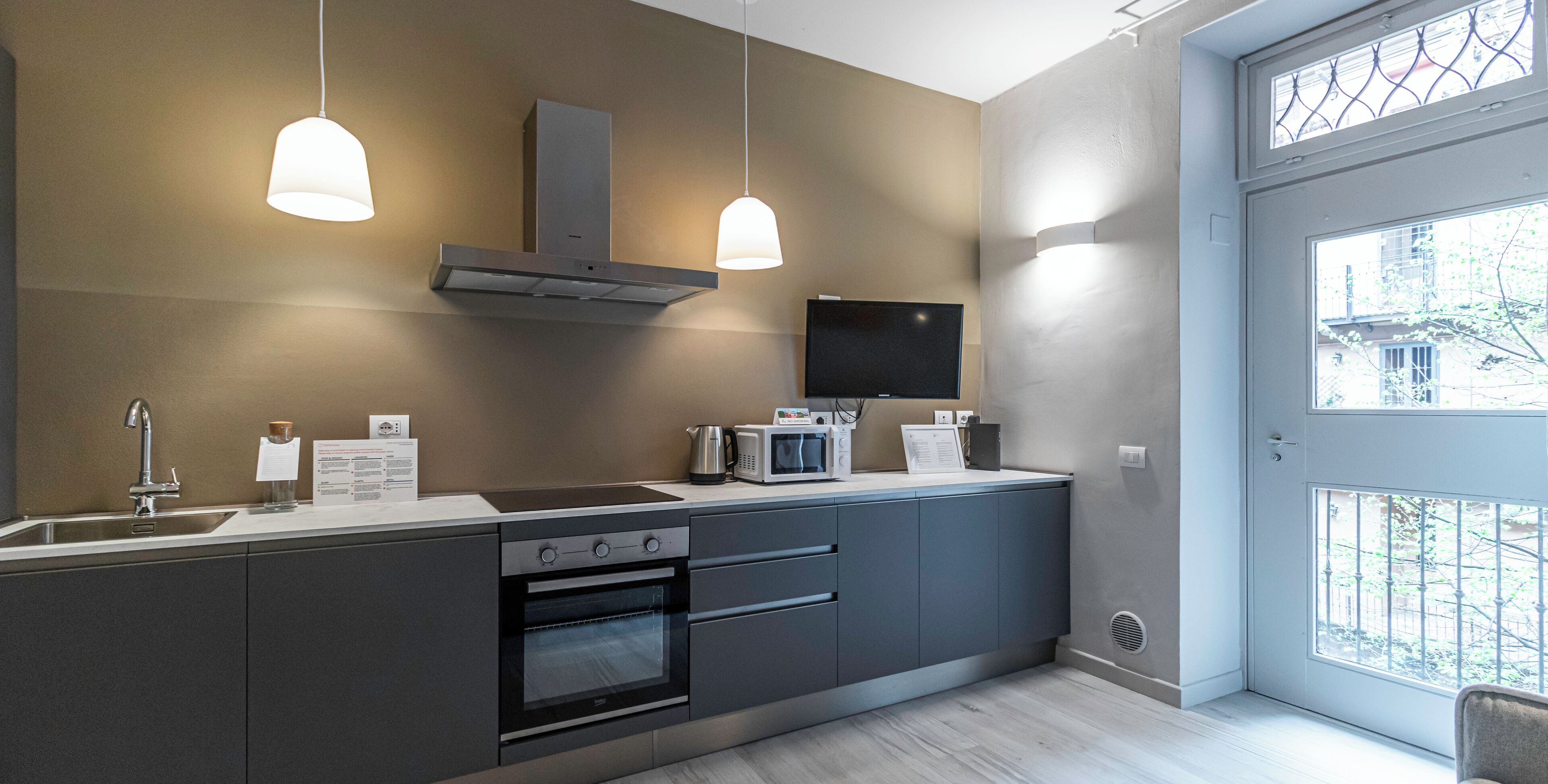 A modern kitchen with a built-in oven, microwave, and stainless steel sink. There are pendant lights and a flat-screen TV mounted on the wall.