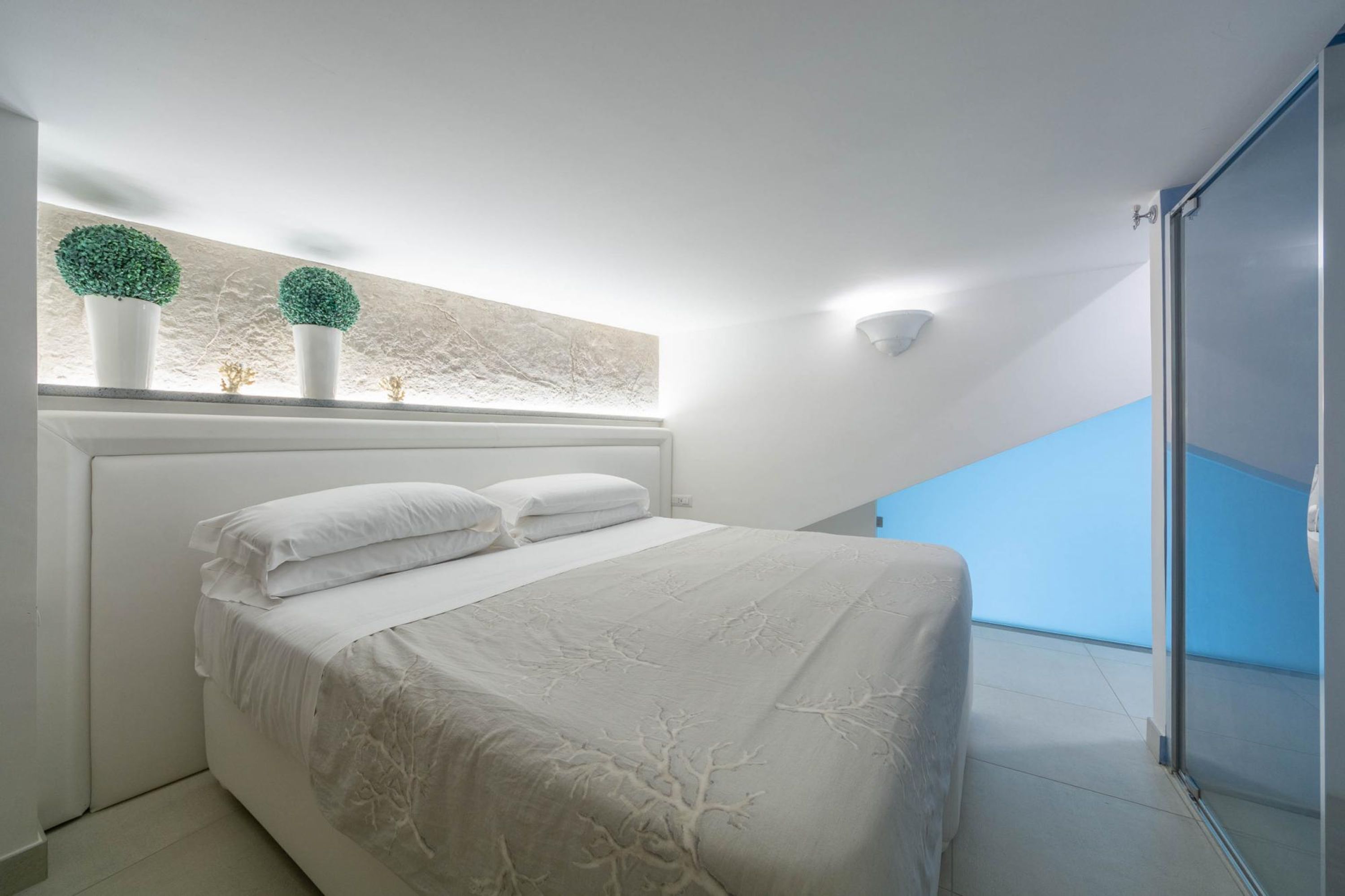 A bedroom with a bed, white bedding, a window with two potted plants, and a view of a balcony.