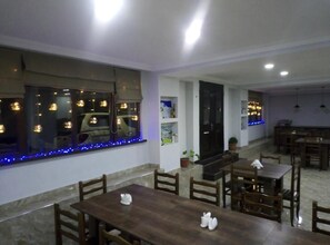 A dining area with wooden tables and chairs, a large window with blue lights, and a kitchen area in the background. - Hotel Gera (Mestia)