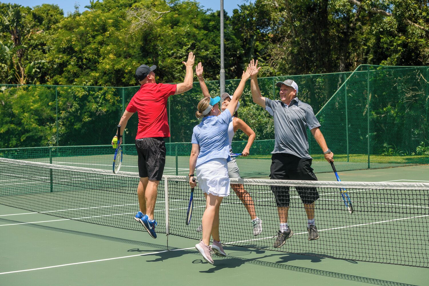 A tennis court with a net, surrounded by a green fence and trees.