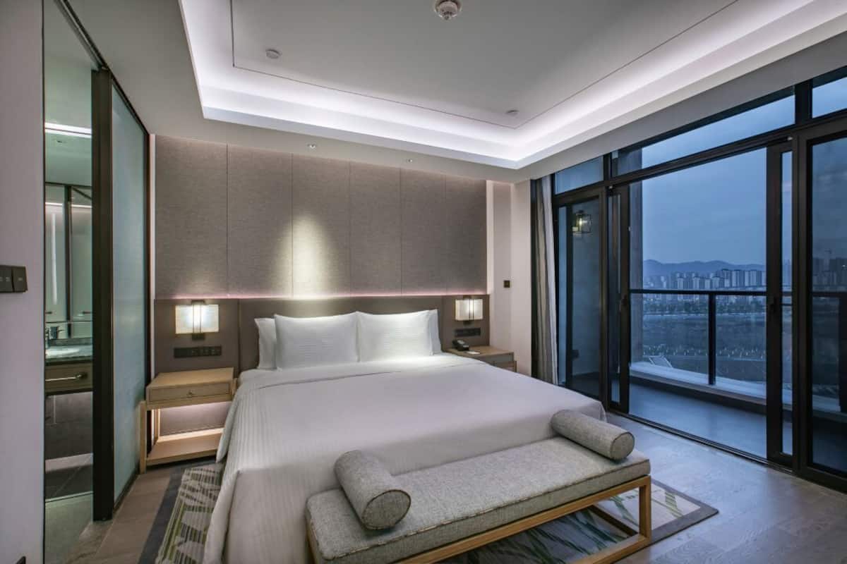 A modern hotel room with a large bed, bedside lamps, a nightstand, a bench, and a large window offering a city view.