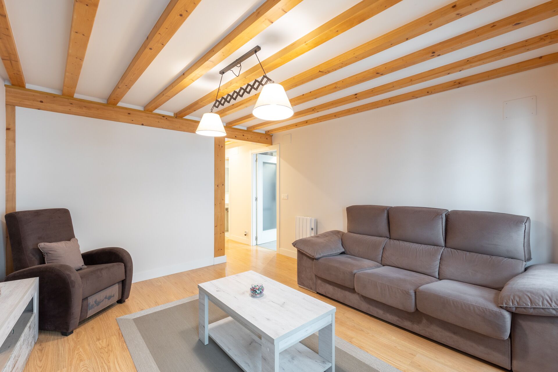 Un salón amplio con techo de madera, un sofá marrón, una mesa de centro blanca y un sillón marrón.