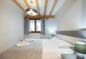 A bedroom with a bed, a nightstand, a lamp, a window with curtains, and a wooden ceiling beam. - Gamarra - baskeyrentals (Lekeitio)