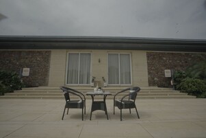 Un área de estar al aire libre con dos sillas y una mesa, un muro de piedra y la entrada de un edificio con escalones.