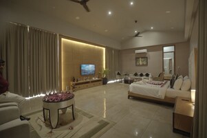 Quarto de hotel moderno com uma cama grande, área de estar com mesa de centro, televisão e uma janela ampla com cortinas.