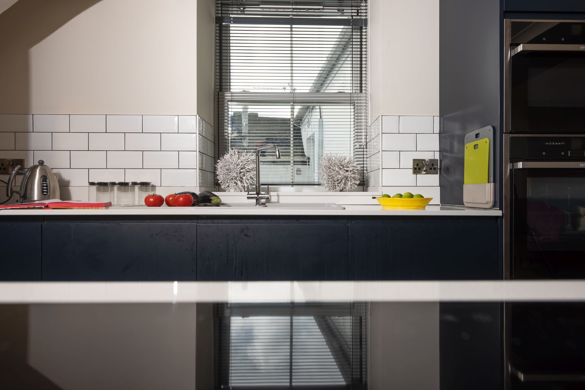 Una cocina moderna con encimera blanca, electrodomésticos de acero inoxidable y una ventana con persianas.