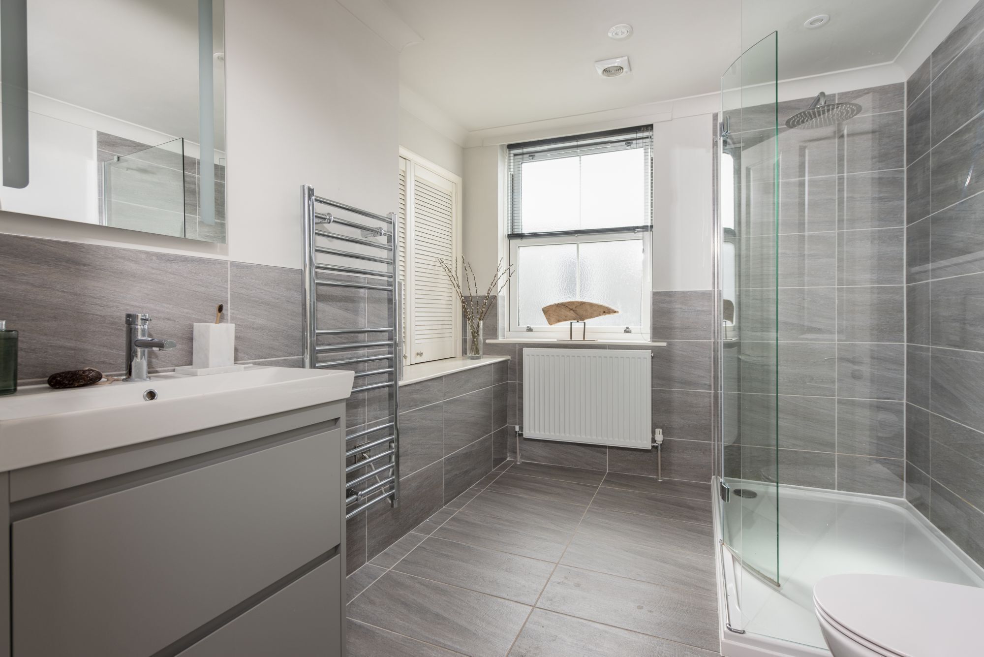 A modern bathroom with a glass shower enclosure, a white sink, and a window with blinds.