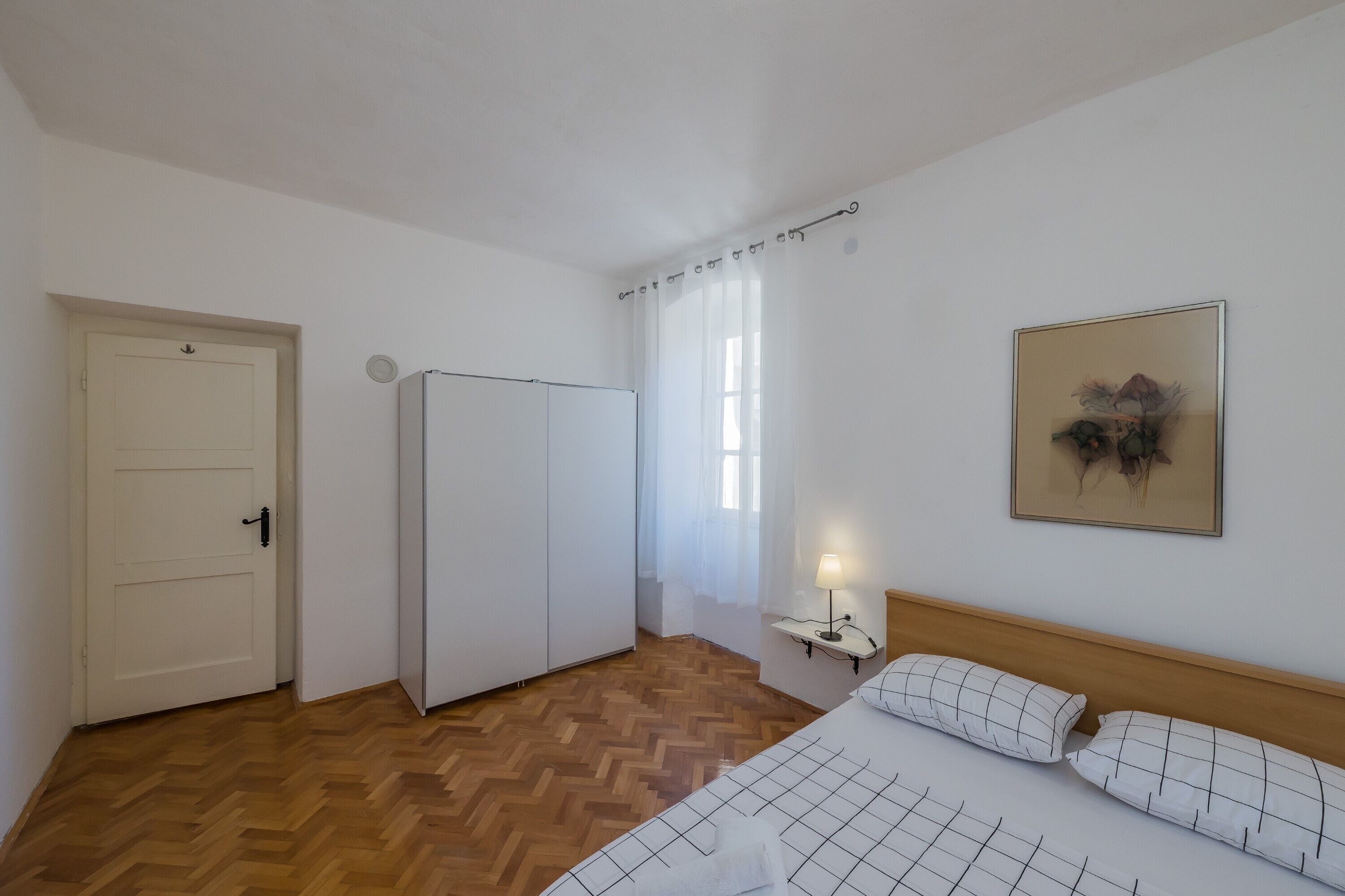 Ein Hotelzimmer mit Holzboden, einem Bett mit weißer Bettwäsche, einem Nachttisch mit Lampe, einer Tür und einem Fenster mit durchsichtigen Vorhängen.