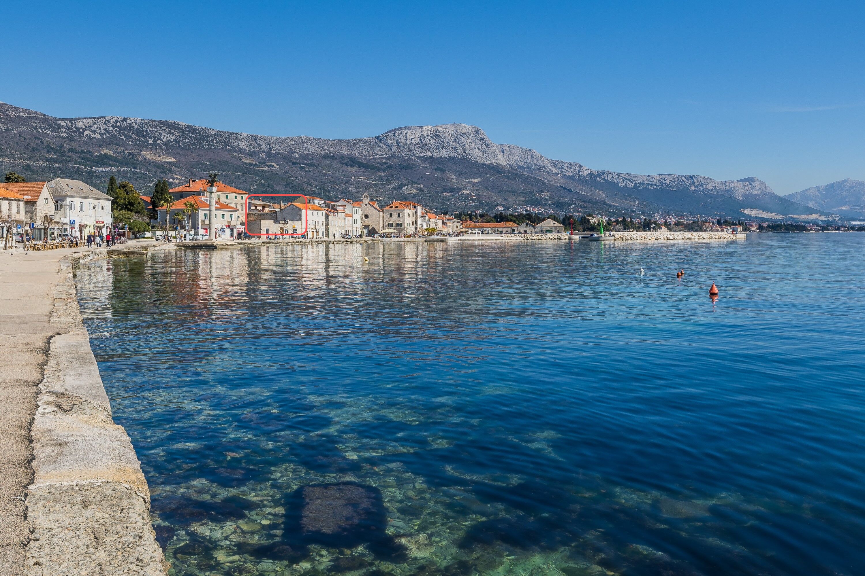 A waterfront view with clear water, buildings, and mountains in the background.